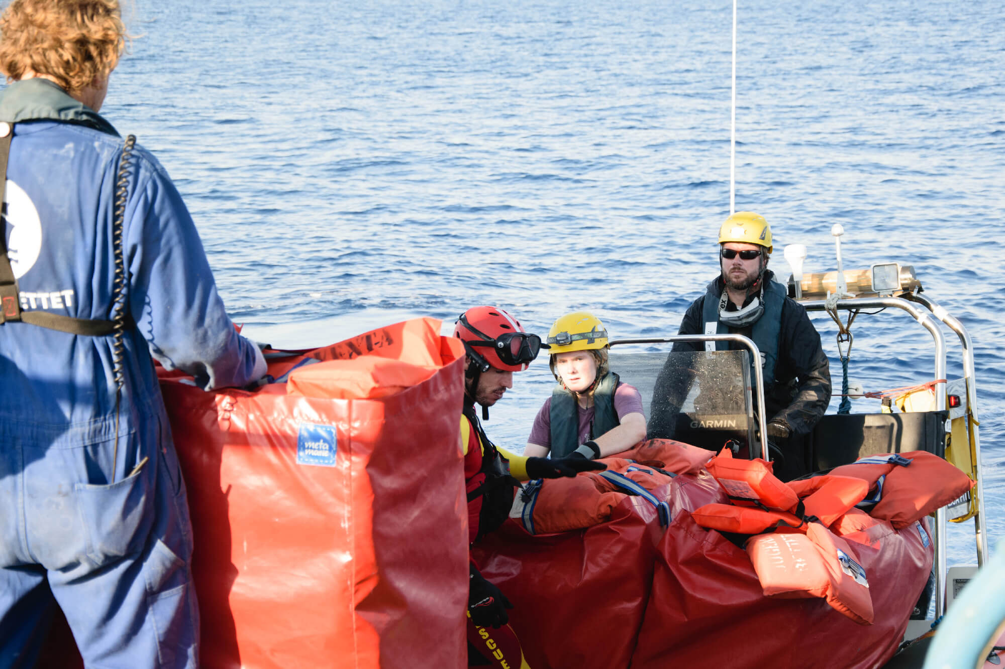 The Ocean in the Background, in the Foreground a person with red buybacks filled with rescue-jackets which are handed over to crew-members on a RIB.